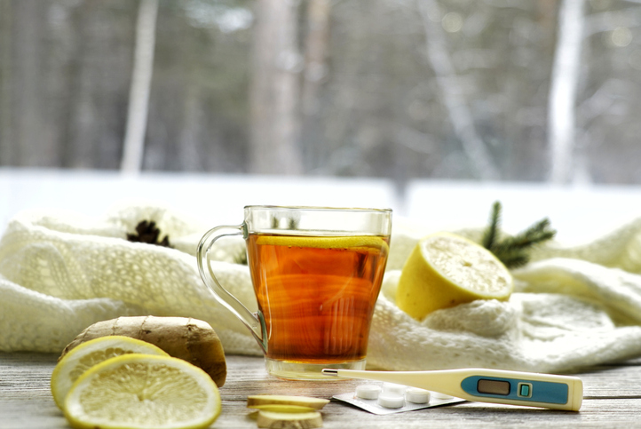 Glazen kopje thee met citroen- en gemberwortelschijfjes, muntblaadjes op een glazen schoteltje op een rustieke houten tafel met op de achtergrond de wollen wanten en gebreide sjaal van de vrouwen buiten. koorts behandelen